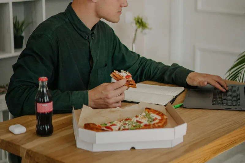 Mann isst am Laptop Pizza und trinkt Cola – Stress, schnelles Essen und Verdauung