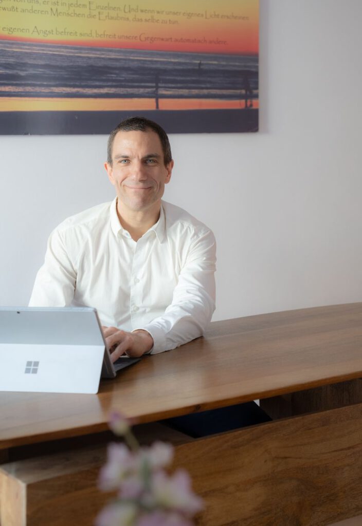 Heilpraktiker Harald Erb lächelnd am Schreibtisch mit Laptop – transparente Kosten für Darmgesundheit, Praxis Berlin Prenzlauer Berg
