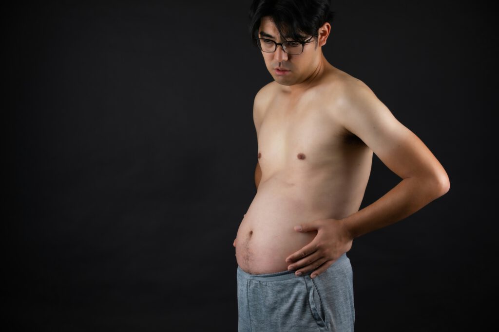 Mann mit nacktem Oberkörper schaut auf seinen vollen, vorgewölbten Unterbauch – Völlegefühl nach dem Essen